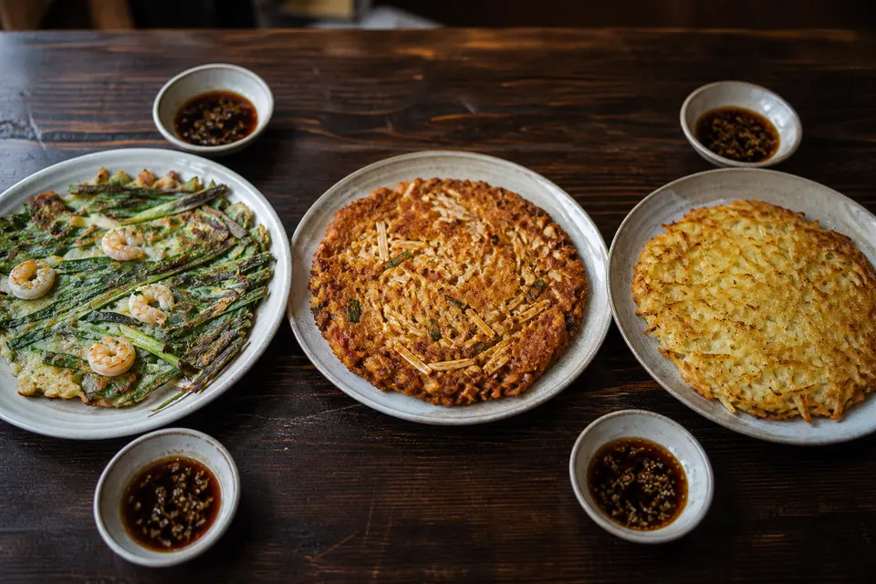 Three hyperreal Korean savory pancakes arranged for comparison: scallion pajeon, mung-bean bindaetteok, and potato gamjajeon.