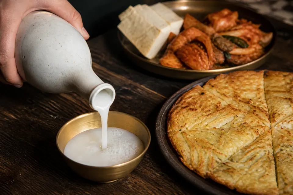 Makgeolli poured into a brass bowl beside sliced potato pancake, tofu, and stir-fried kimchi.
