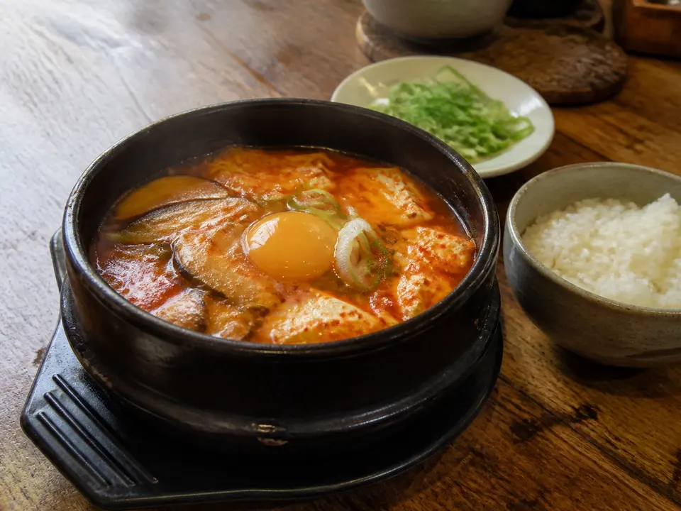 A bubbling sundubu jjigae with soft tofu, egg, mushrooms, scallions, and a bowl of rice on a wooden table.